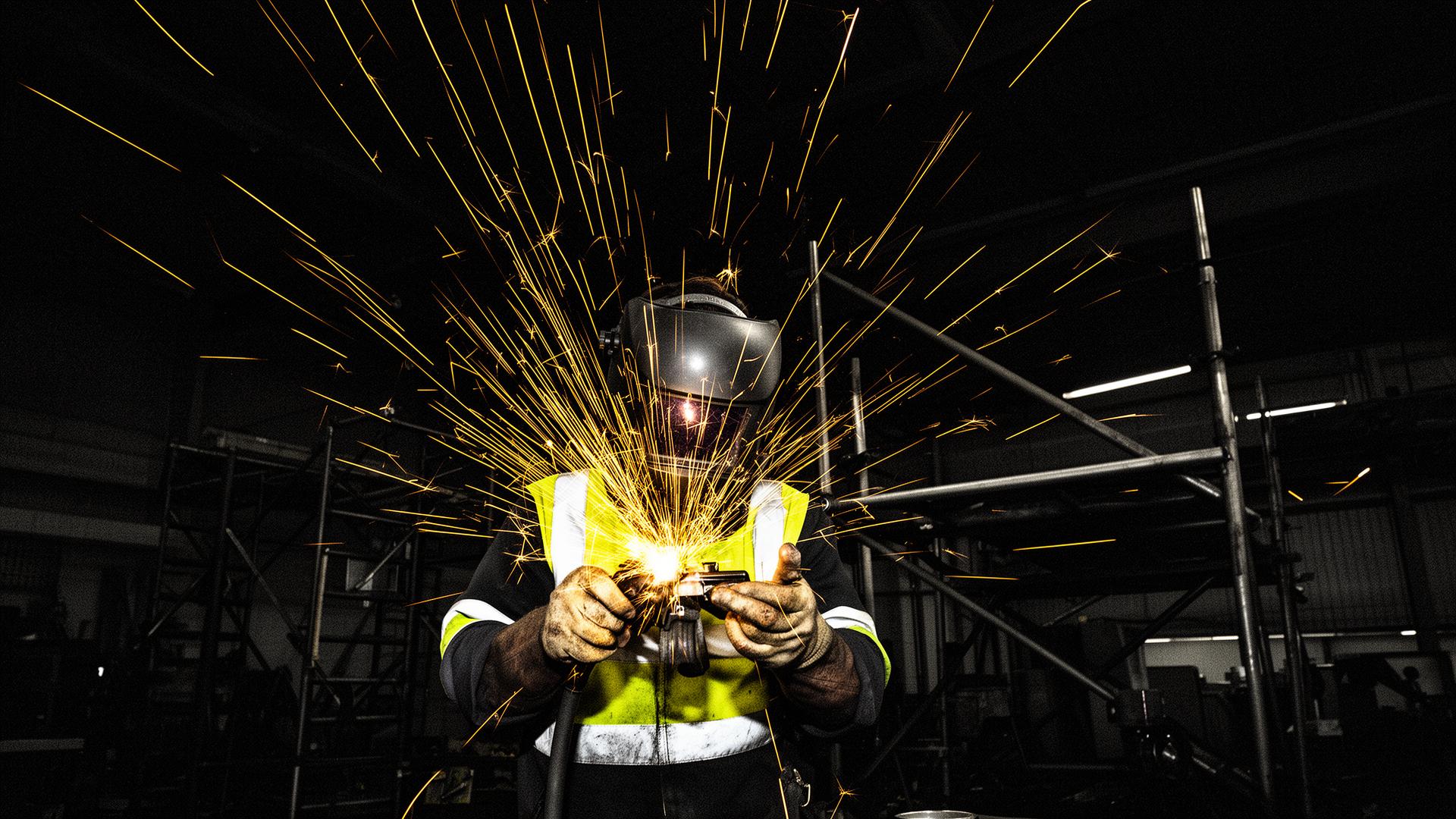 Welder mid-arc, sparks showering against a dark industrial workshop